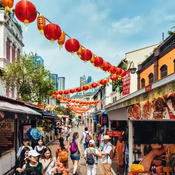 Red lanterns hang over a narrow street filled with pedestrians shopping at market stalls in a city.