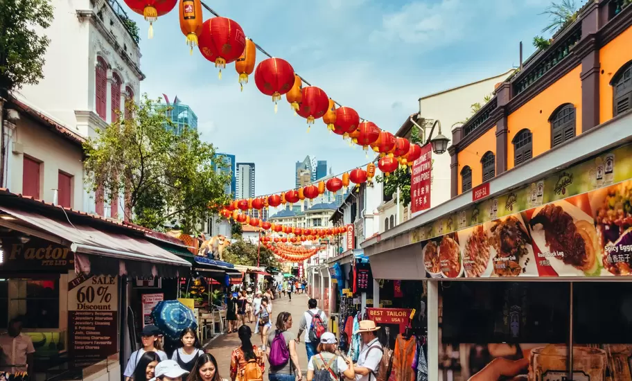 Red lanterns hang over a narrow street filled with pedestrians shopping at market stalls in a city.