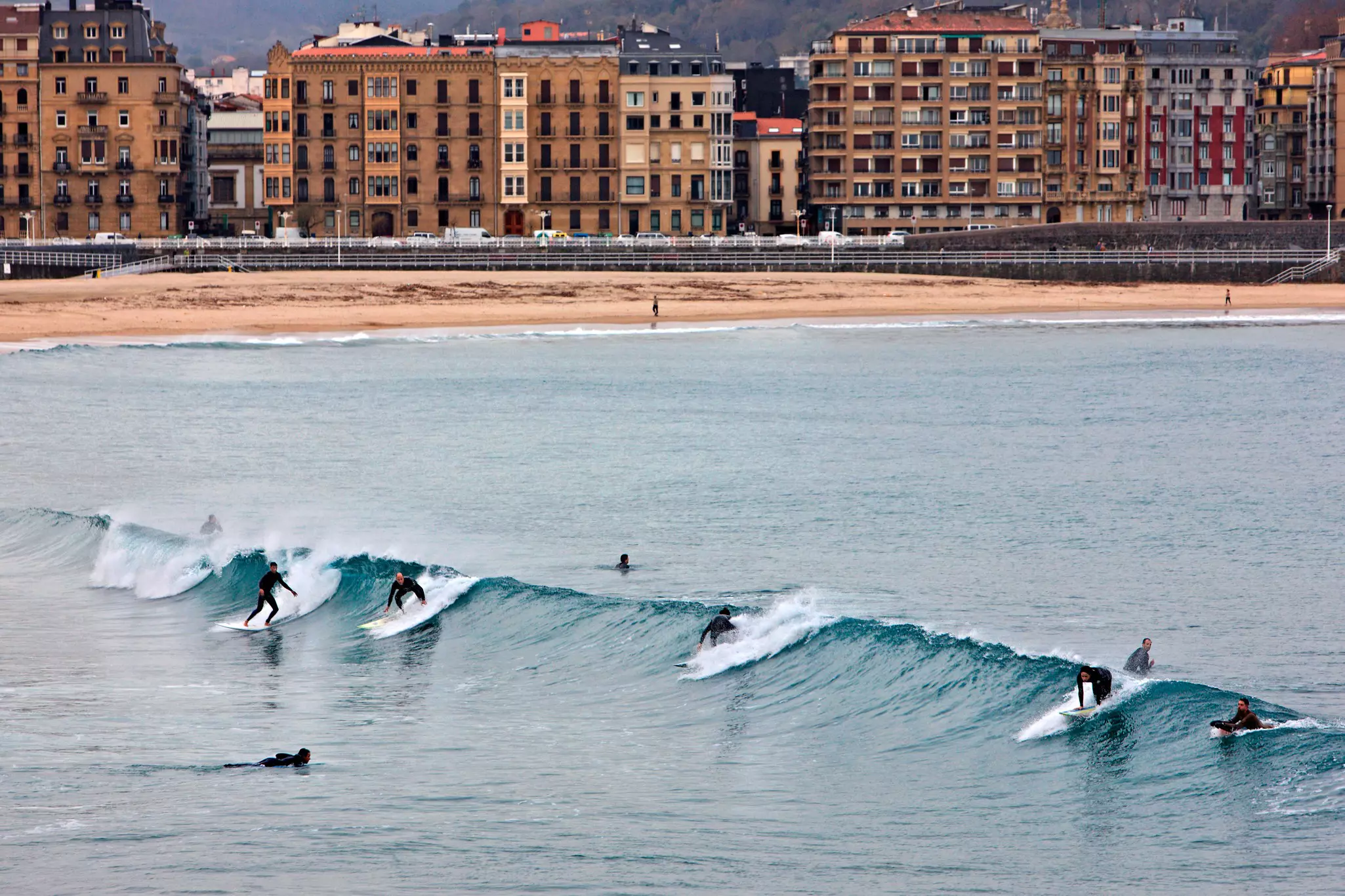 Surfers in wetusits catch a wave in the ocean near a city beach.