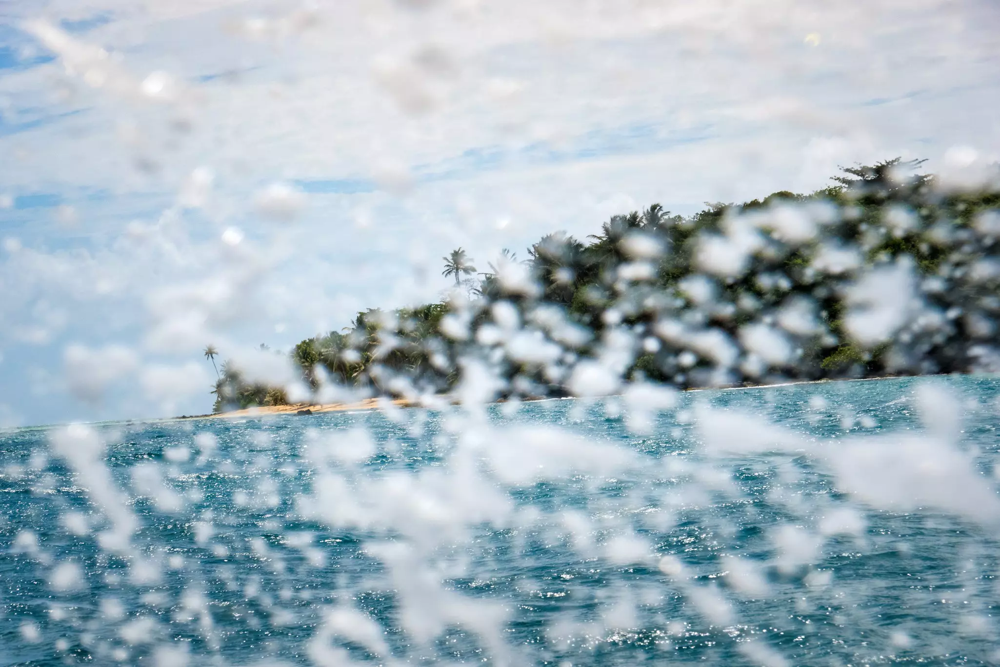 Water splashes up from the edge of a boat as it travels towards an island covered in palm trees.