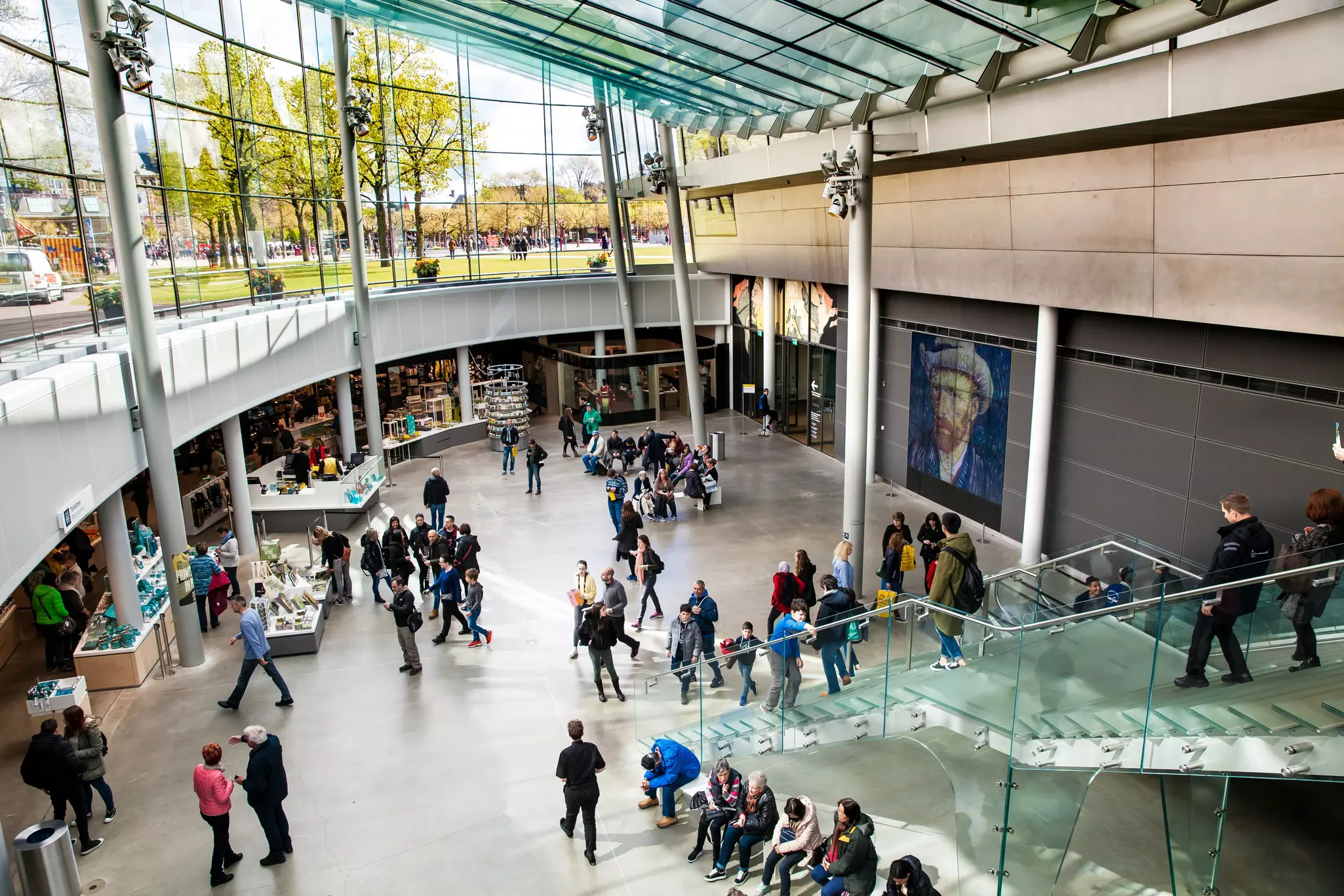 Bird's-eye view of people walking and resting in a museum lobby, with glass-sided staircase to the right, shop to the left, and a painting of Vincent Van Gogh on the righthand wall. Glass windows above show a grassy outdoor scene.