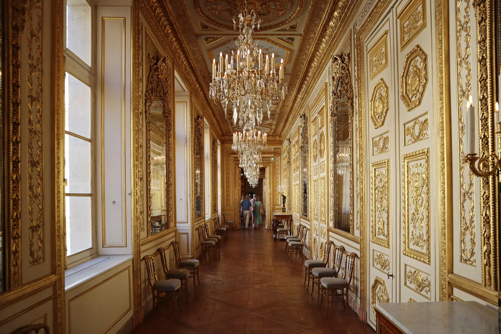 A corridor with gold features and details on the woodwork, around the doors and windows. Large chandeliers hang from the ceiling at intervals.