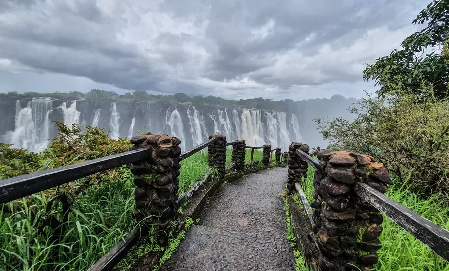 Victoria Falls: a cobbled path with stone-pillar railings leads toward the wide curtain of falling water under storm clouds