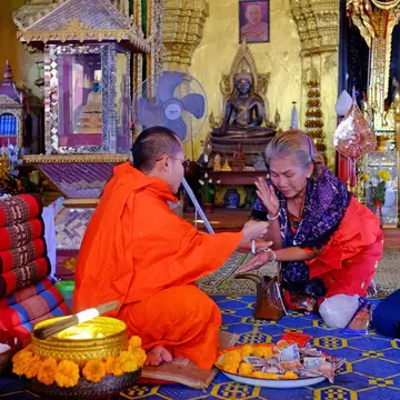 A monk performing a blessing before a bronze Buddha, worshippers kneeling with offerings.