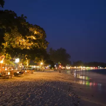 Night on a Sihanoukville beach, Cambodia: rattan chairs and lanterns under a lit tree, bar lights curving along the shoreline