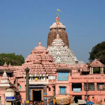 Crowds gather before the pink gateway and tall stone spire of the Jagannath Temple under a clear blue sky in Puri.