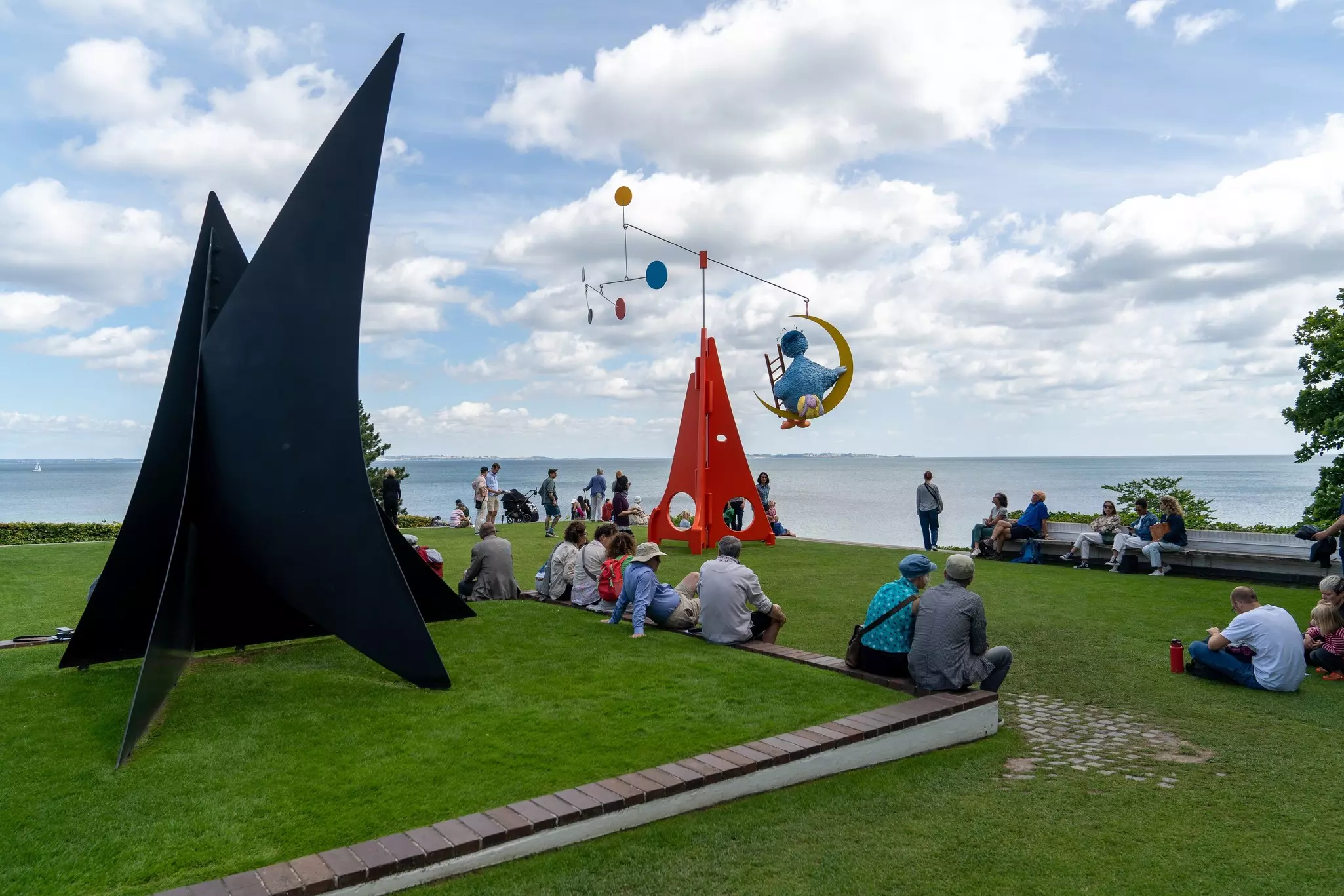People sitting and standing on a green lawn with large artworks by the water.