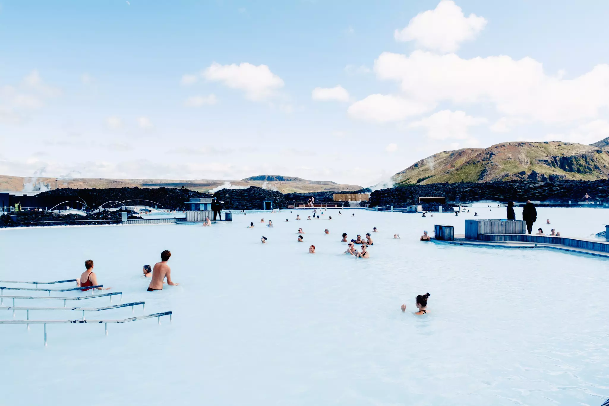 People in a milky blue pool; steam rises from black rocks on the shore.
