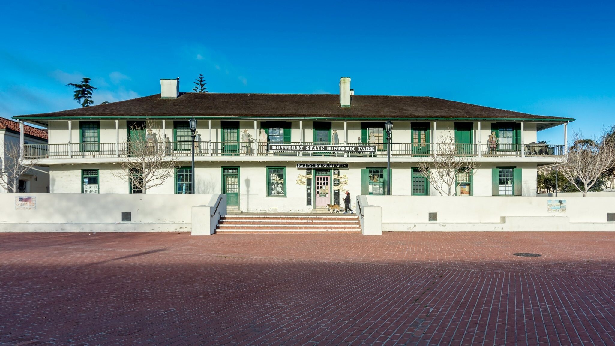 Two-storey whitewashed adobe building with green shutters and a long balcony fronting an empty red-brick plaza under blue sky