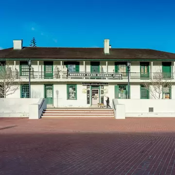 Two-storey whitewashed adobe building with green shutters and a long balcony fronting an empty red-brick plaza under blue sky