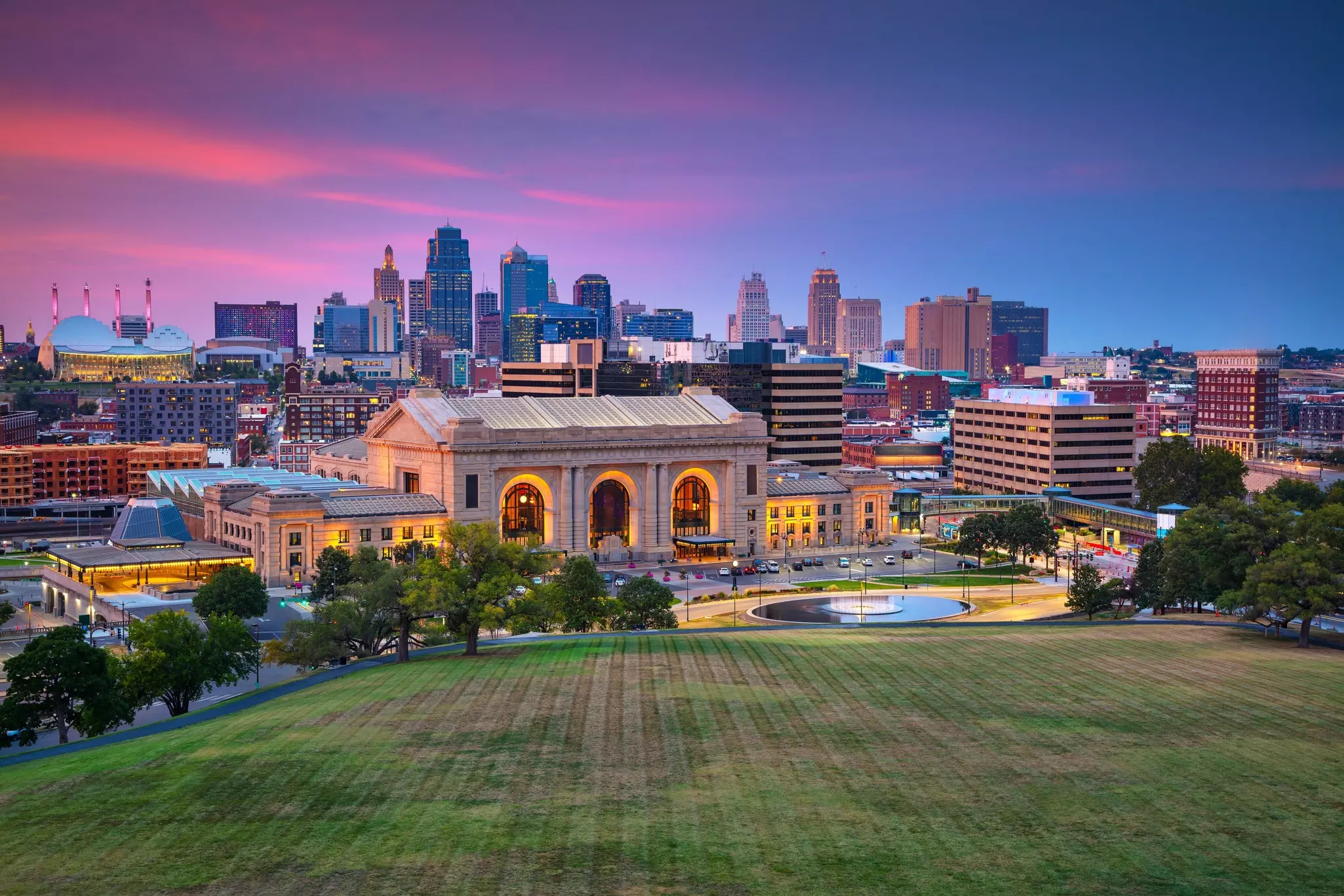 A view of a historic train station and a city skyline beyond at sunset, shot from a hilltop lawn in a park.