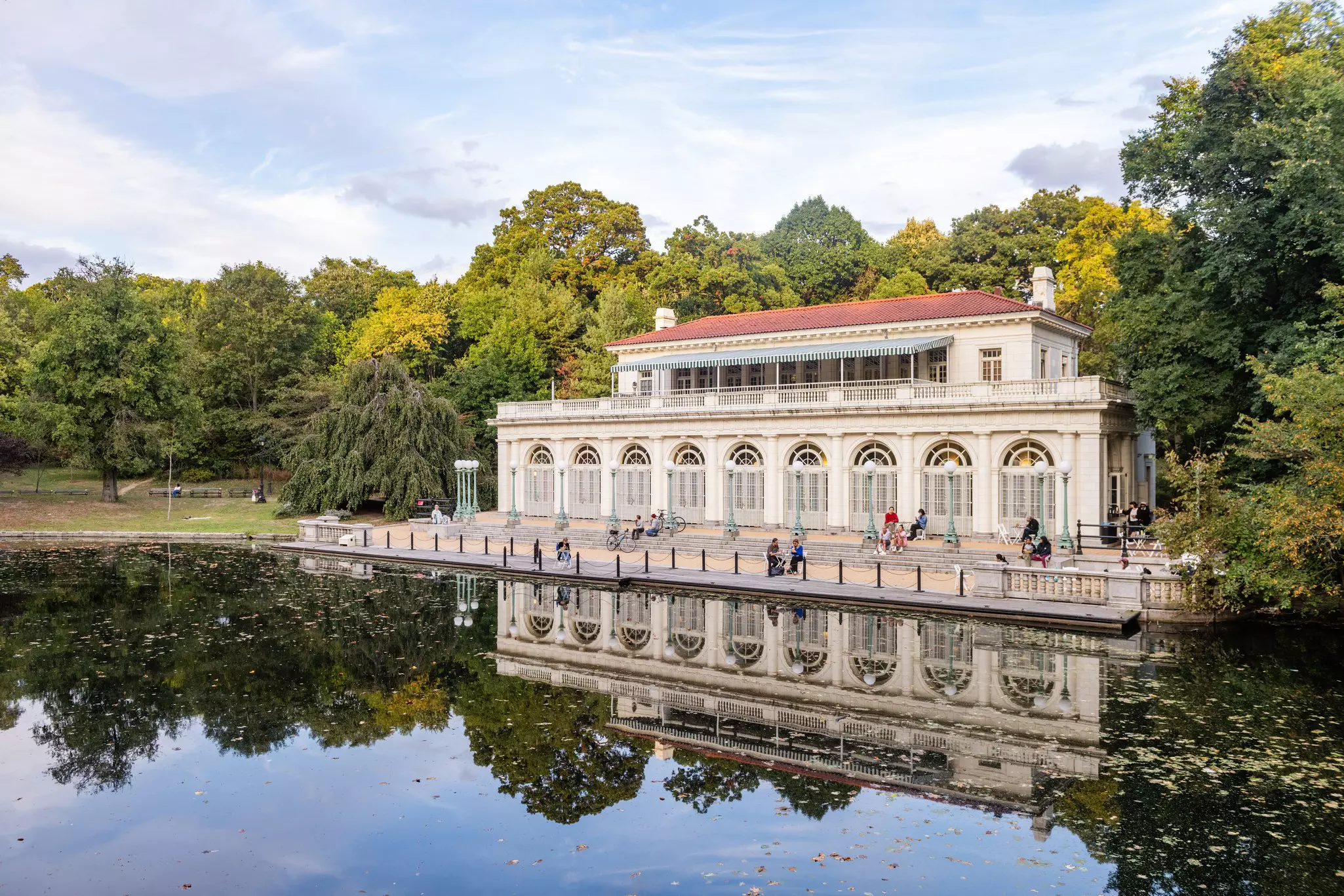 People sit on the terrace beside a lake outside a grand boathouse