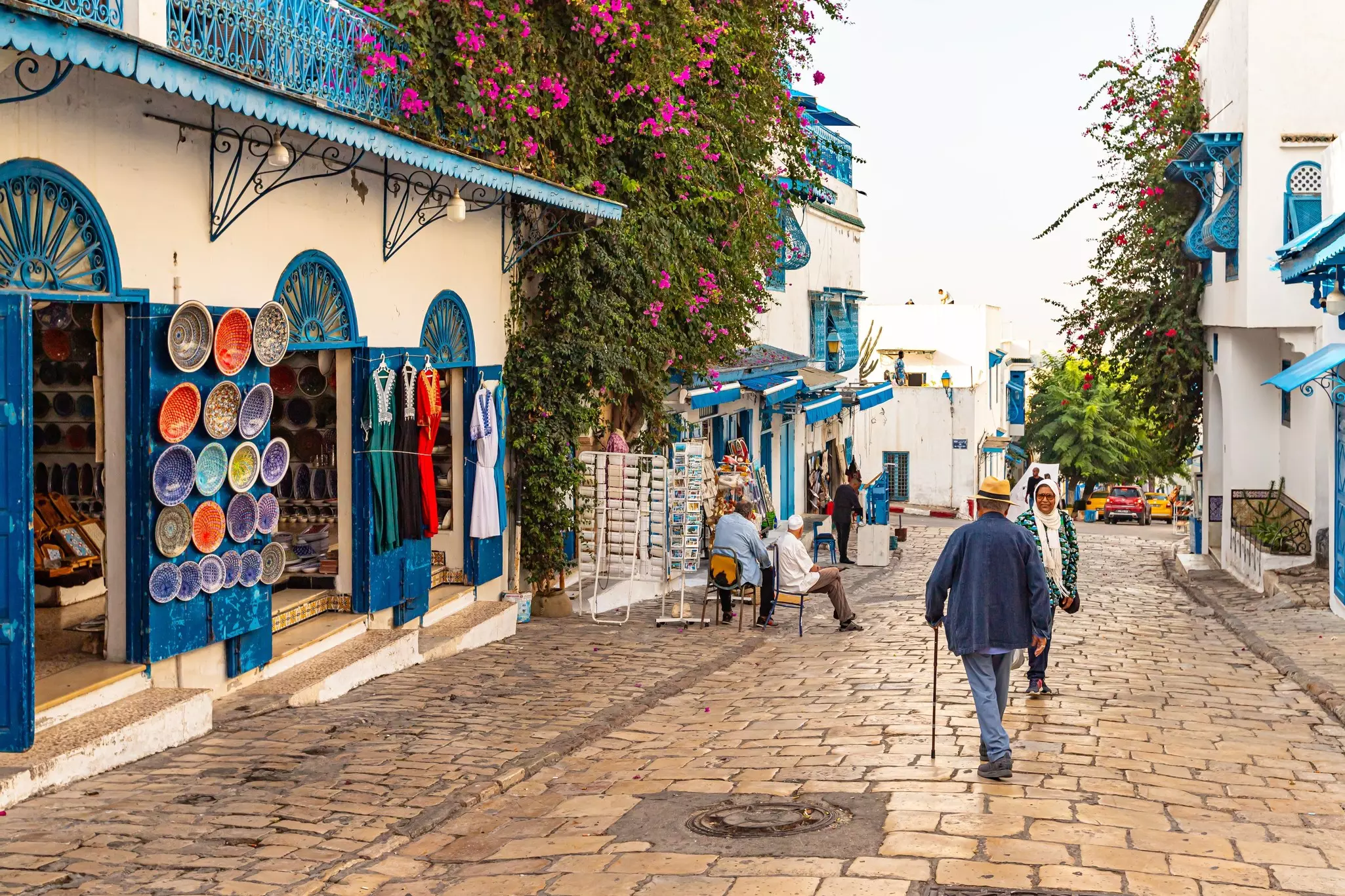 Two people walking in opposite directions on a cobblestone street with white stone buildings painted with turquoise-colored accents and displaying colorful pottery and clothing for sale.