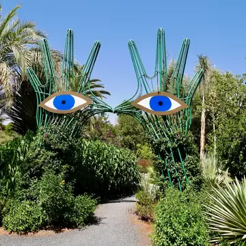 Two green metal hands holding blue-and-white eyes form an arch over a gravel path among palms, shrubs and agave