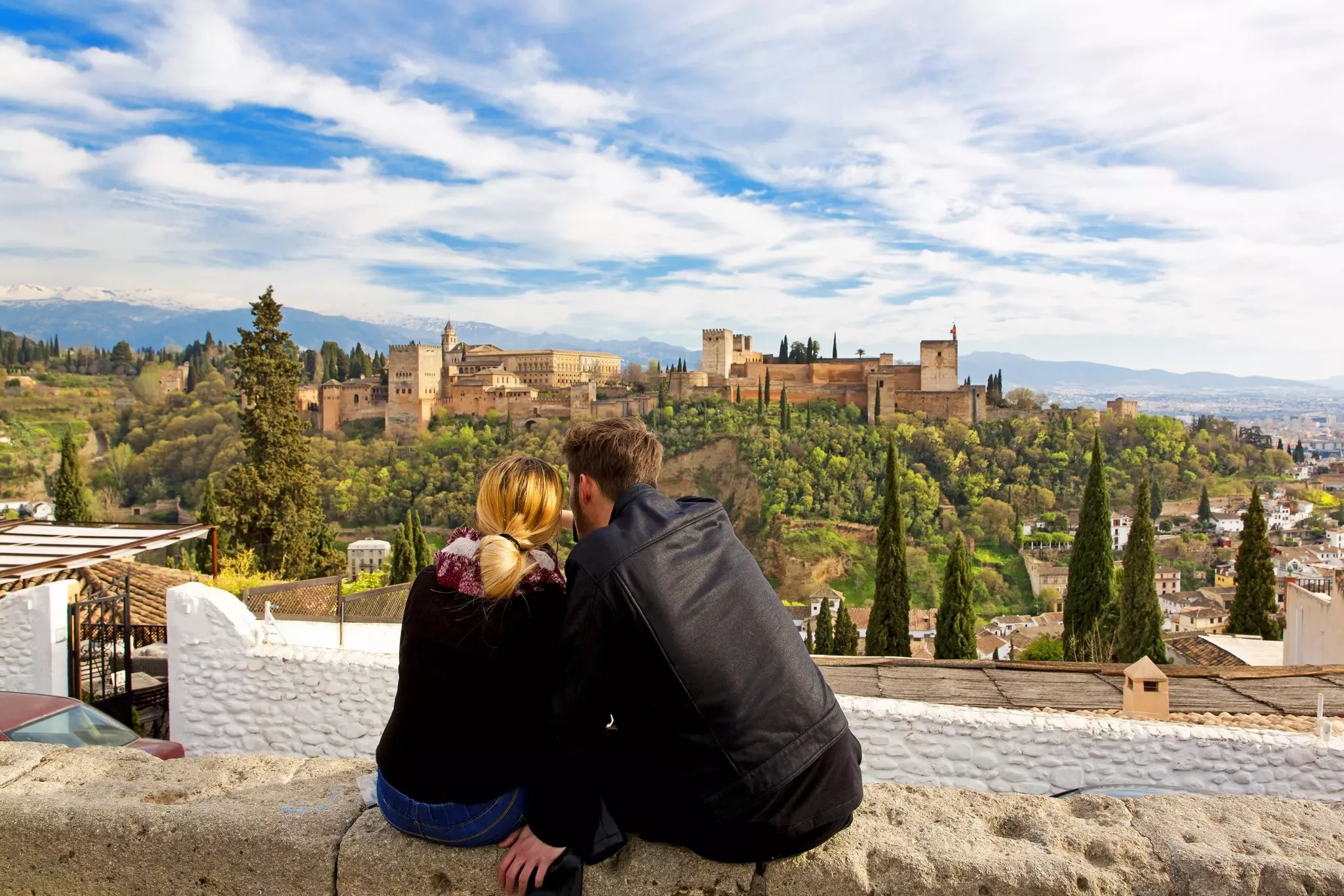 A couple wearing black jackets sit on a wall overlooking Granada and the Alhambra in Spain.
