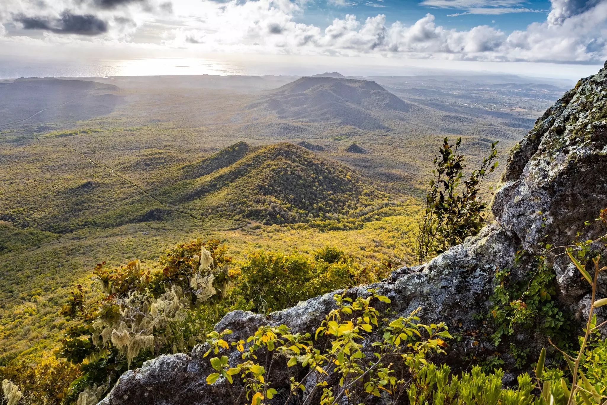 A view of verdant hills from the top of a mountain. Dappled sunbeams illuminate the scene.