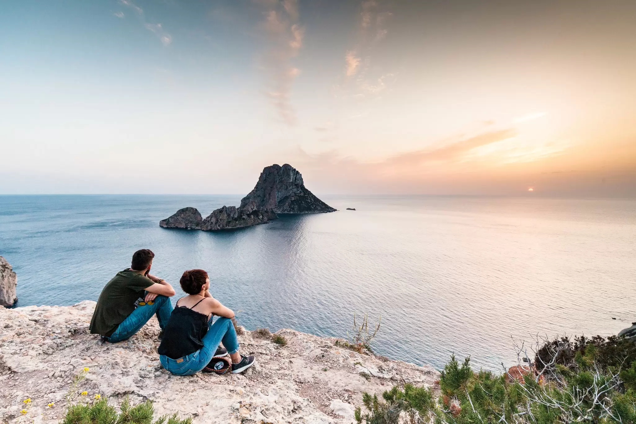 Two people sit on a clifftop watching sunset over the sea in Es Vedrà, Ibiza