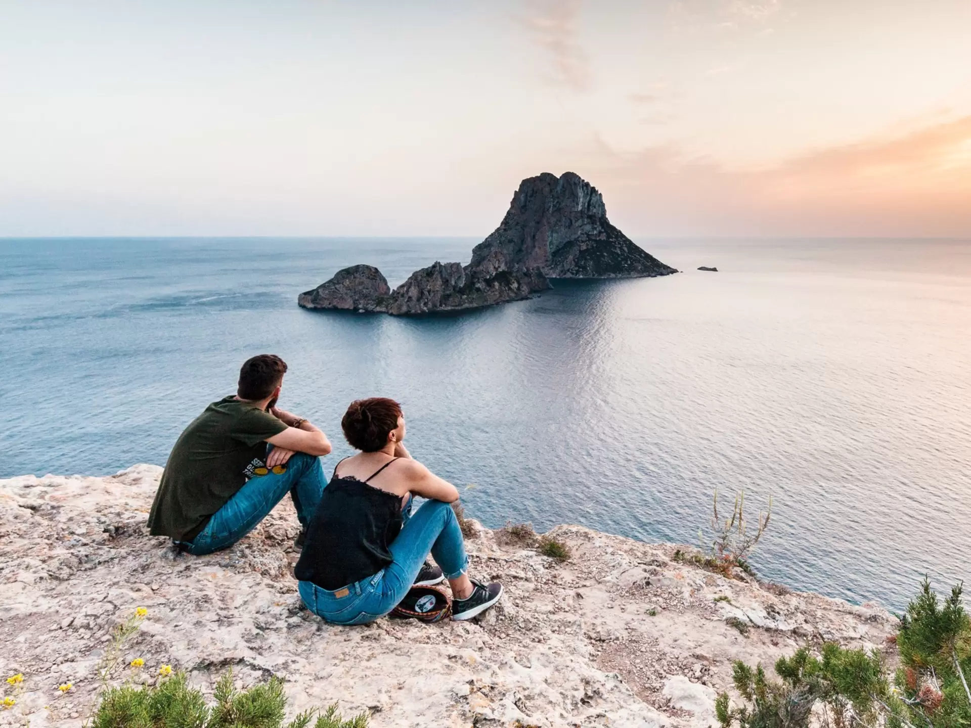 Two people sit on a clifftop watching sunset over the sea in Es Vedrà, Ibiza