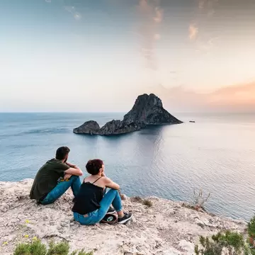 Two people sit on a clifftop watching sunset over the sea in Es Vedrà, Ibiza