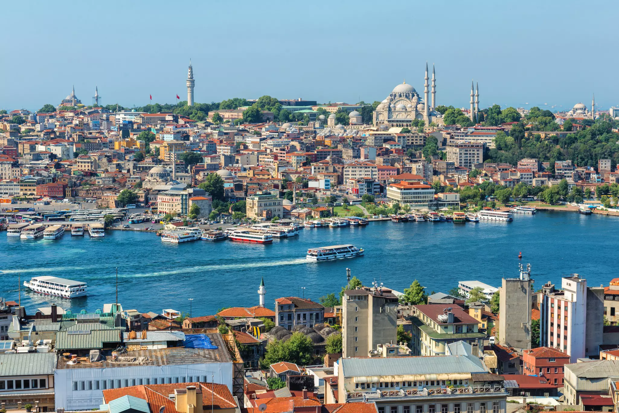 Ferries traveling in a waterway with others docked on a sunny day. City buildings can be seen in the foreground and across the water in the distance.