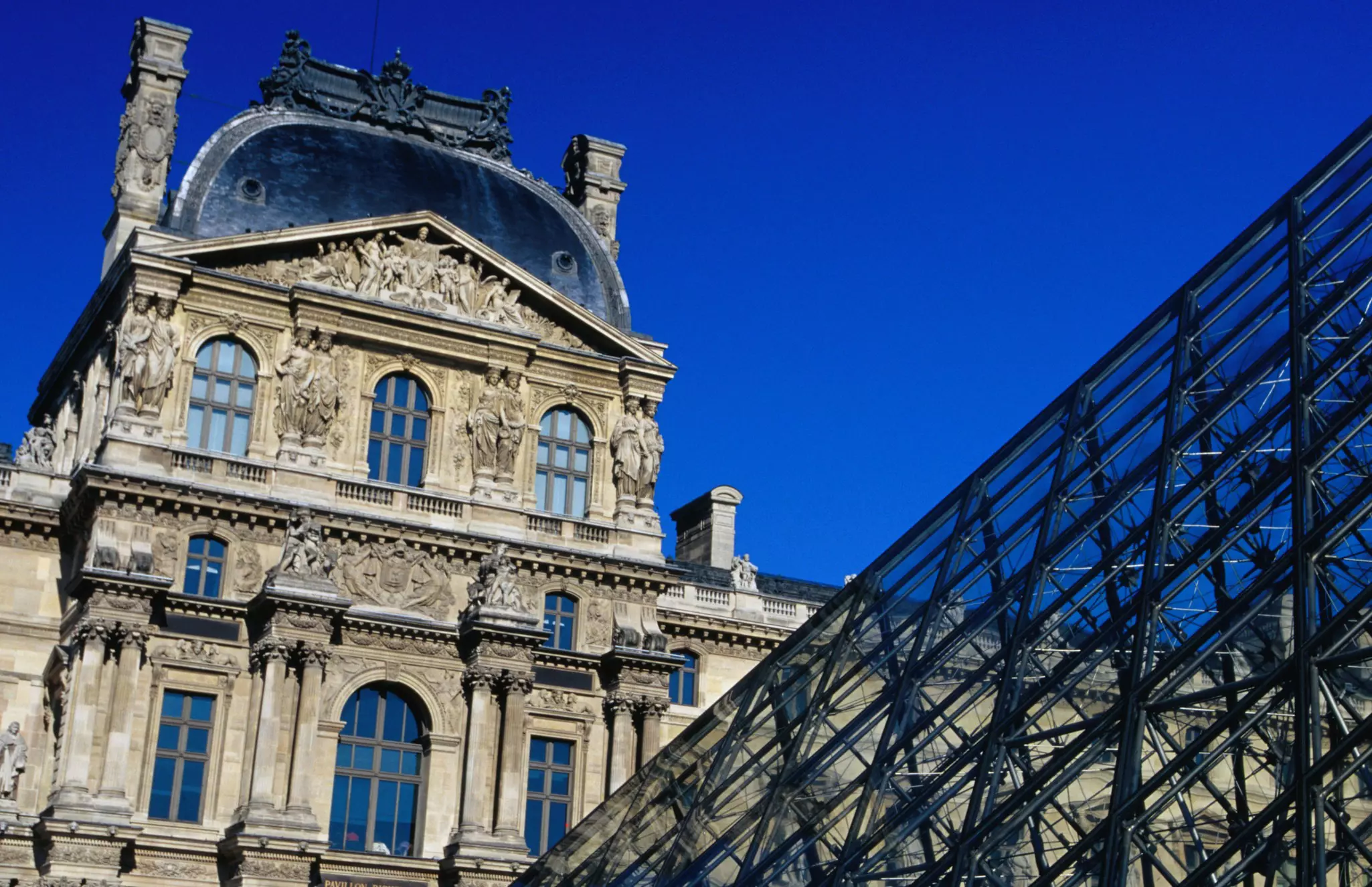 The edge of a glass pyramid against a neoclassical building under bright blue sky in Paris.
