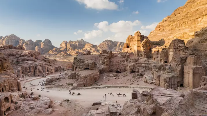 Ancient ruins built into the rock faces of cliffs with camels and people down below. 