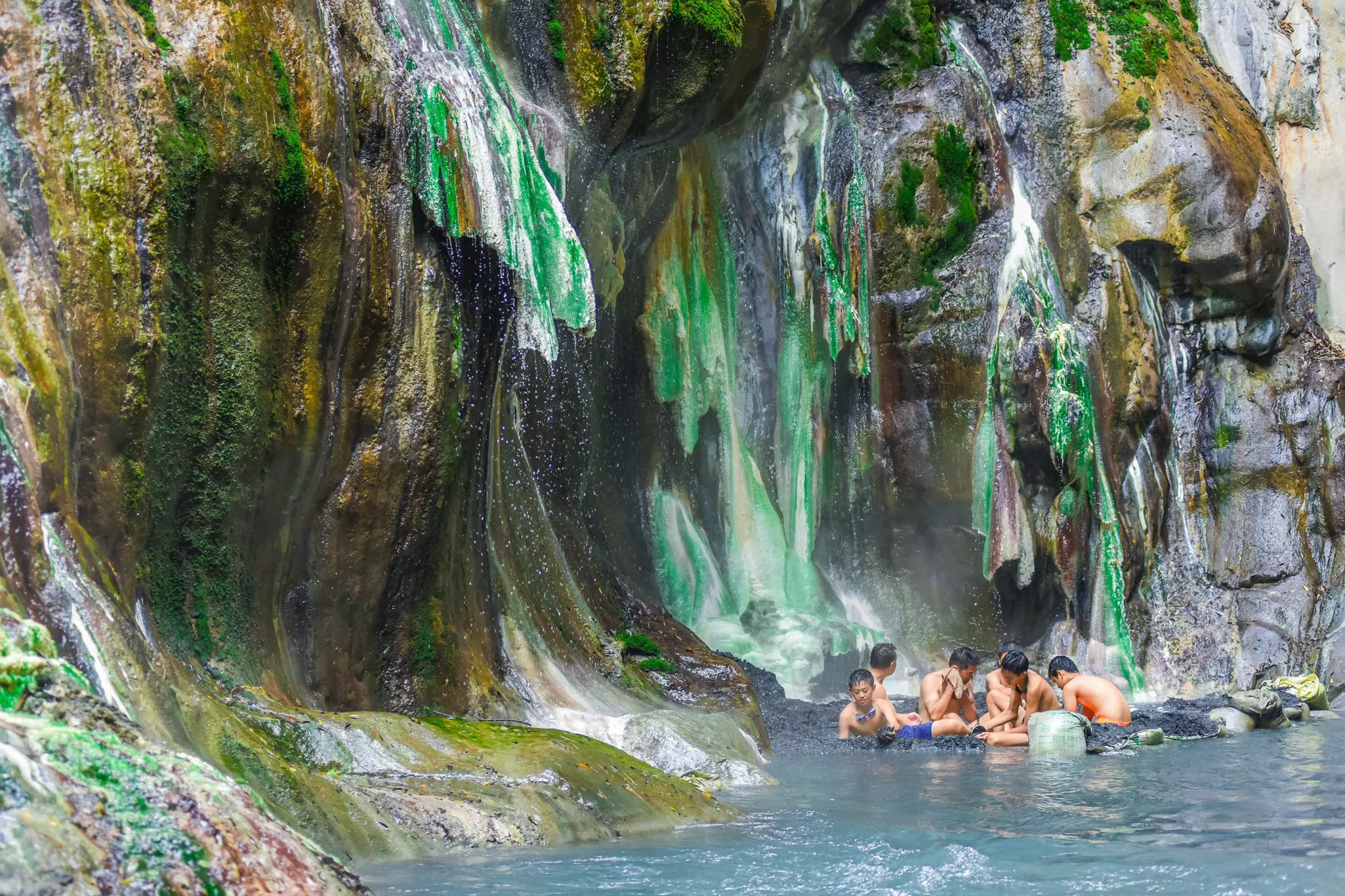 People soak in a hot spring underneath a rock formation with many colors due to mineral deposits.
