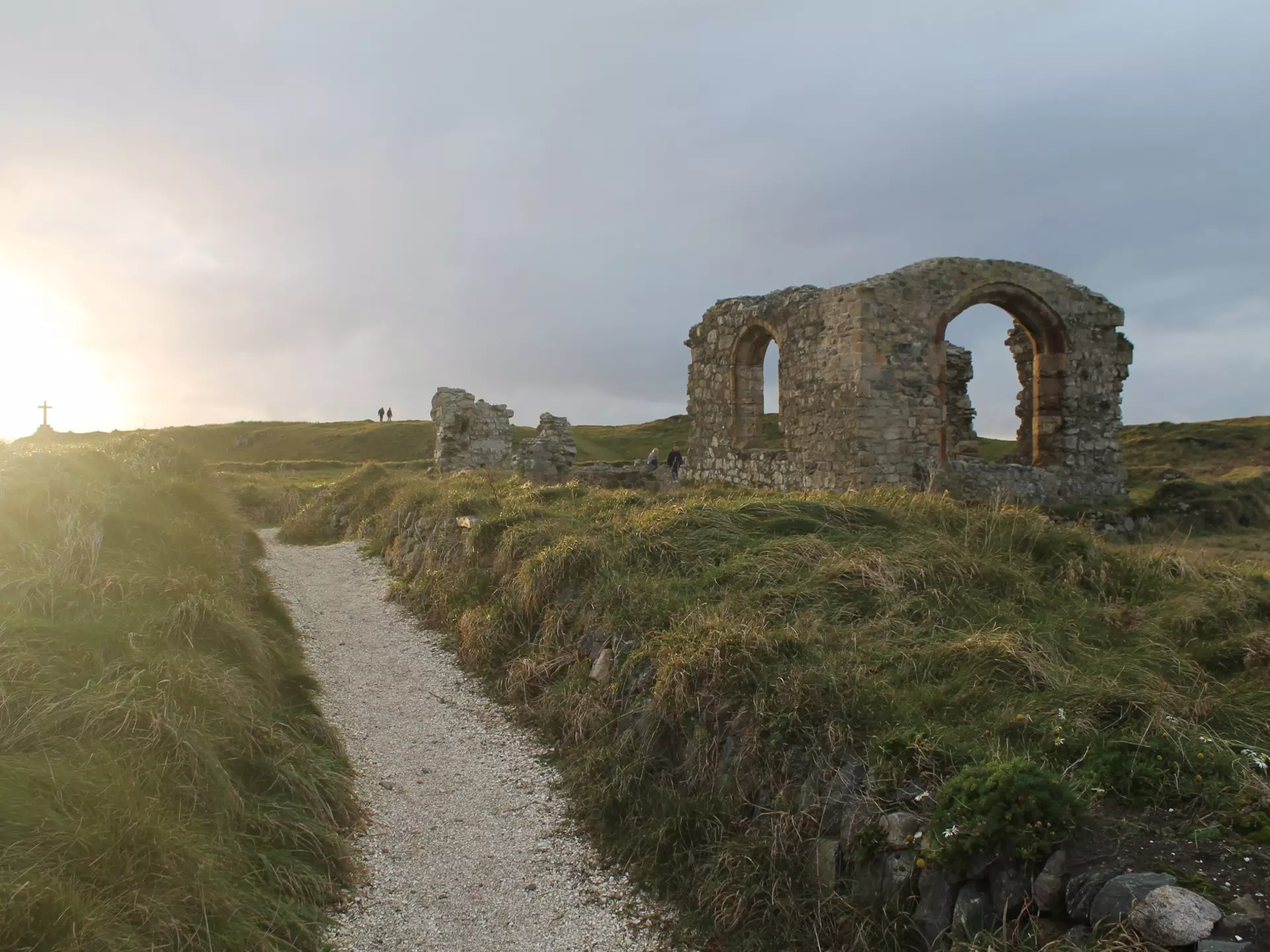 St Dwynwen's Church in Anglesey at sunset. The church is essentially a ruin, with only parts of the walls remaining. Surrounding the church is thick grassland with a narrow walkway to the entrance.