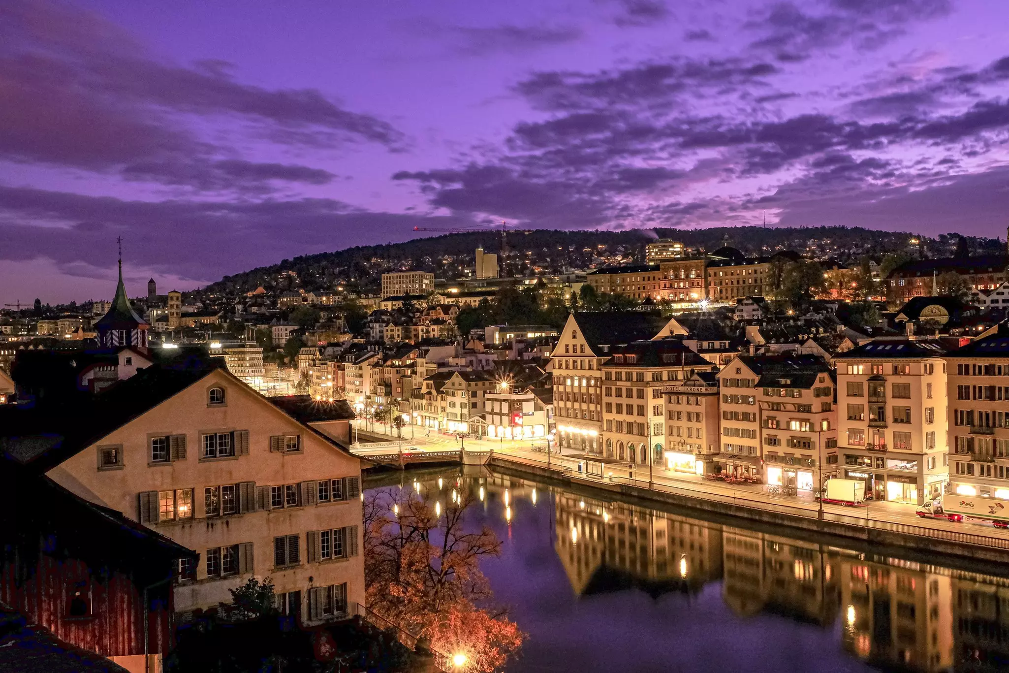 The historic buildings of a city along a river are illuminated at dusk, as pictured from a viewpoint overhead. The evening sky appears purple.