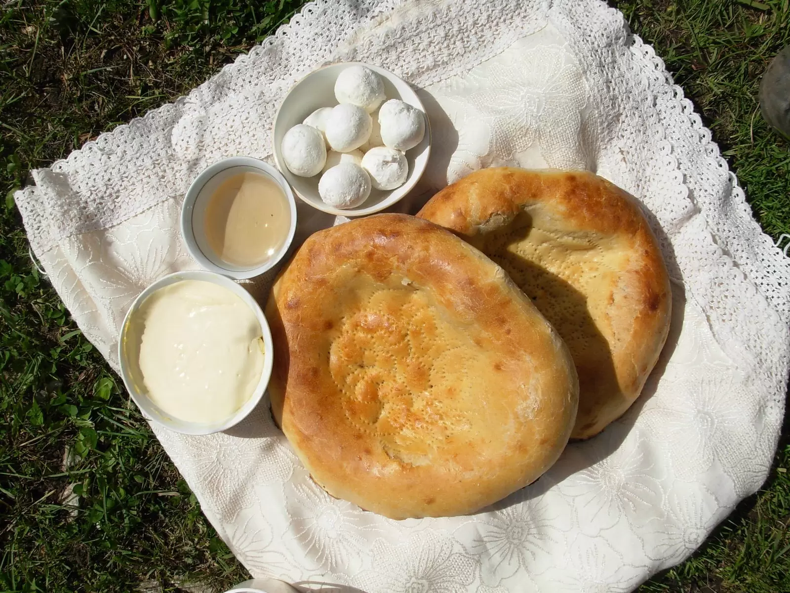 Two round golden flatbread loaves with bowls of cream, butter and white cheese balls on a lace cloth in the grass