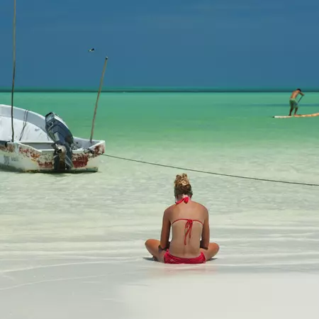 Seascape with boat, paddle board and a young woman reading on the seashore.