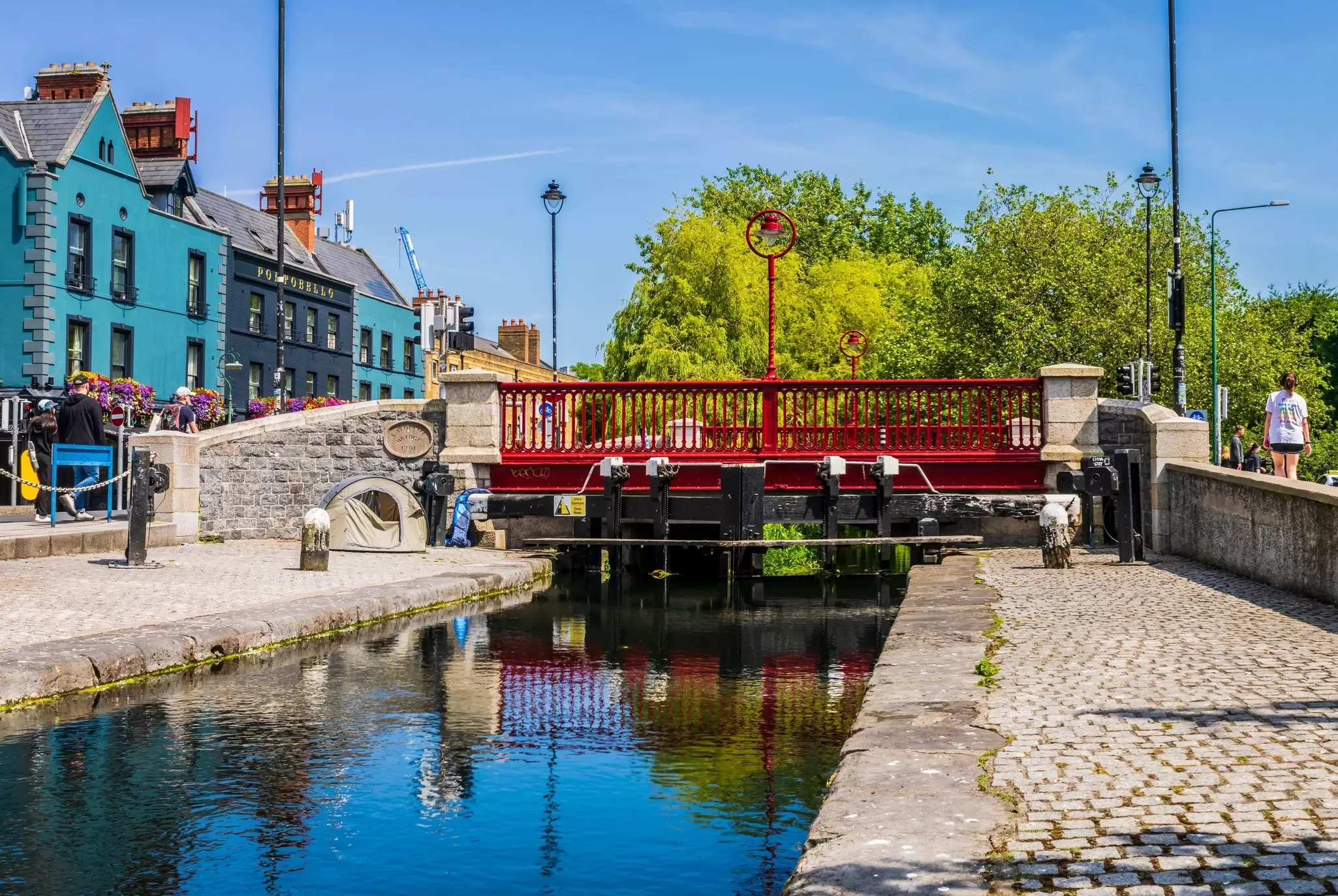 Red bridge spanning a narrow canal bordered by cobblestone with blue buildings in the lefthand background.