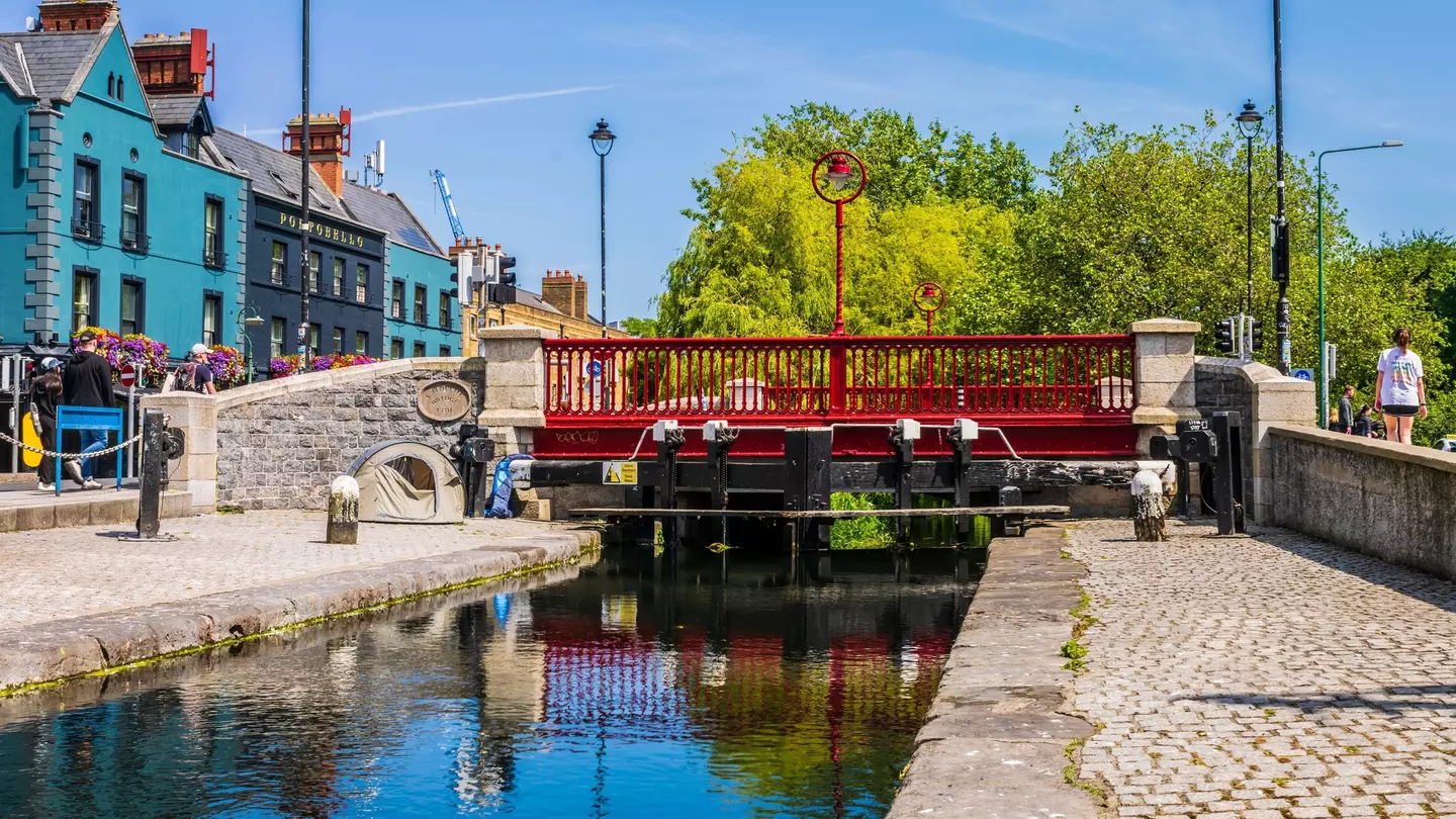 Red bridge spanning a narrow canal bordered by cobblestone with blue buildings in the lefthand background.