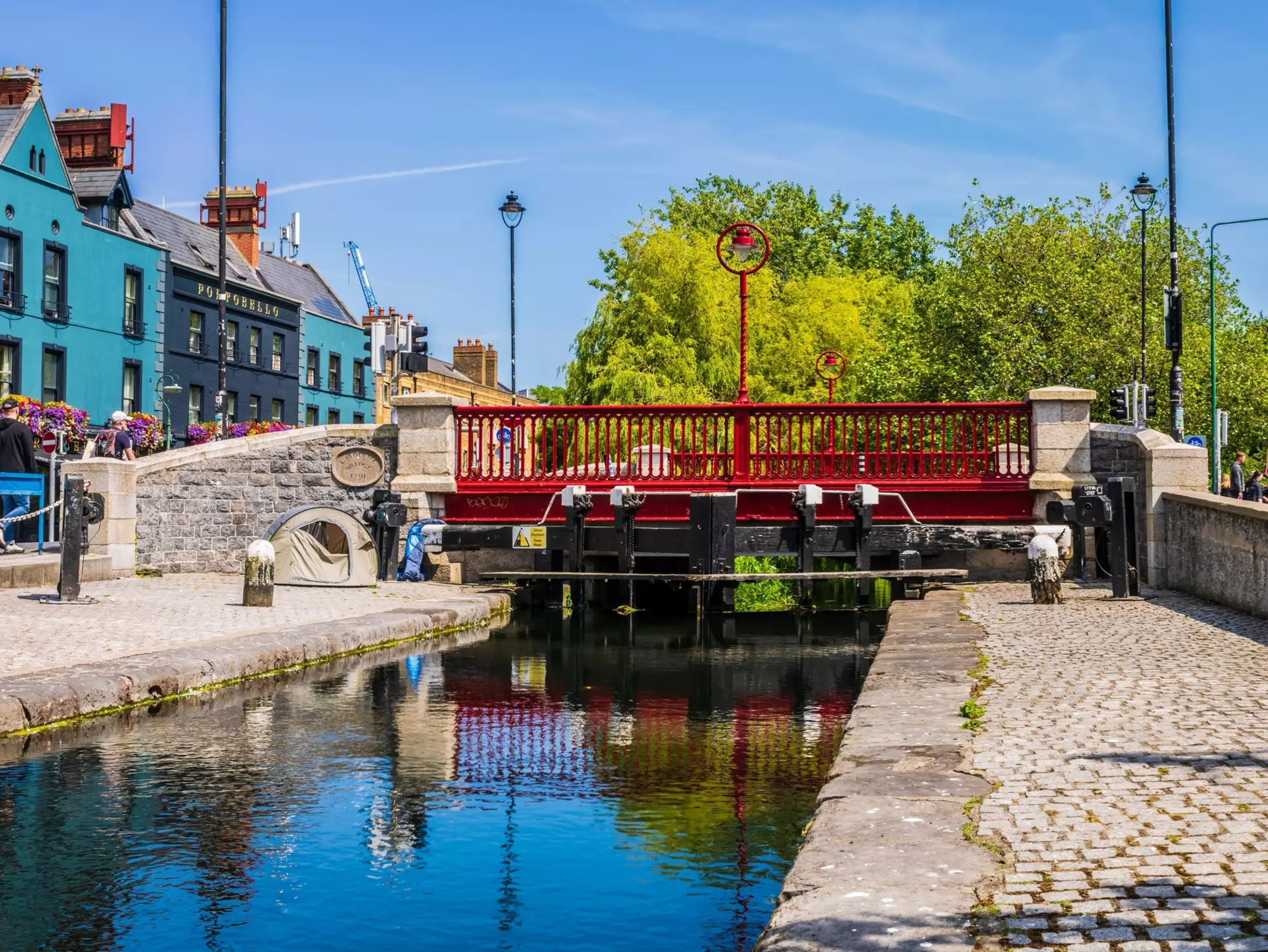 Red bridge spanning a narrow canal bordered by cobblestone with blue buildings in the lefthand background.