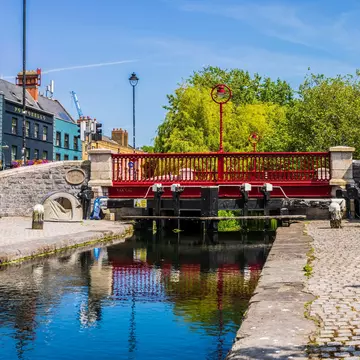 Red bridge spanning a narrow canal bordered by cobblestone with blue buildings in the lefthand background.