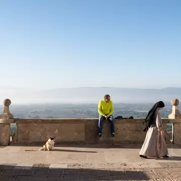 Nun walks across a stone terrace in Assisi as a man and a dog sit by the balustrade above the misty Umbrian plain
