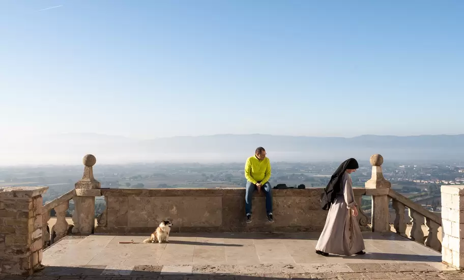Nun walks across a stone terrace in Assisi as a man and a dog sit by the balustrade above the misty Umbrian plain