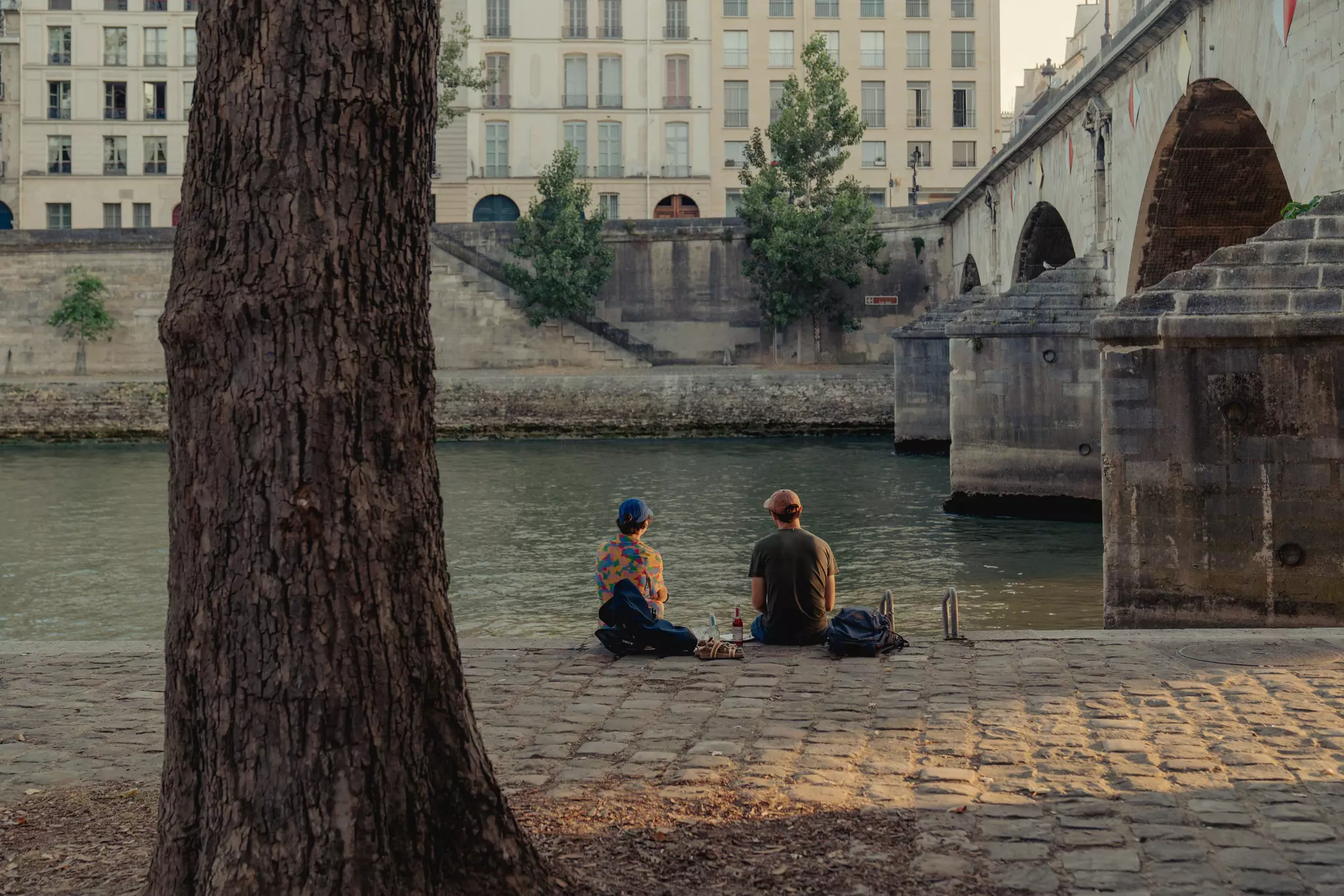 Two people sitting along a river embankment near a bridge