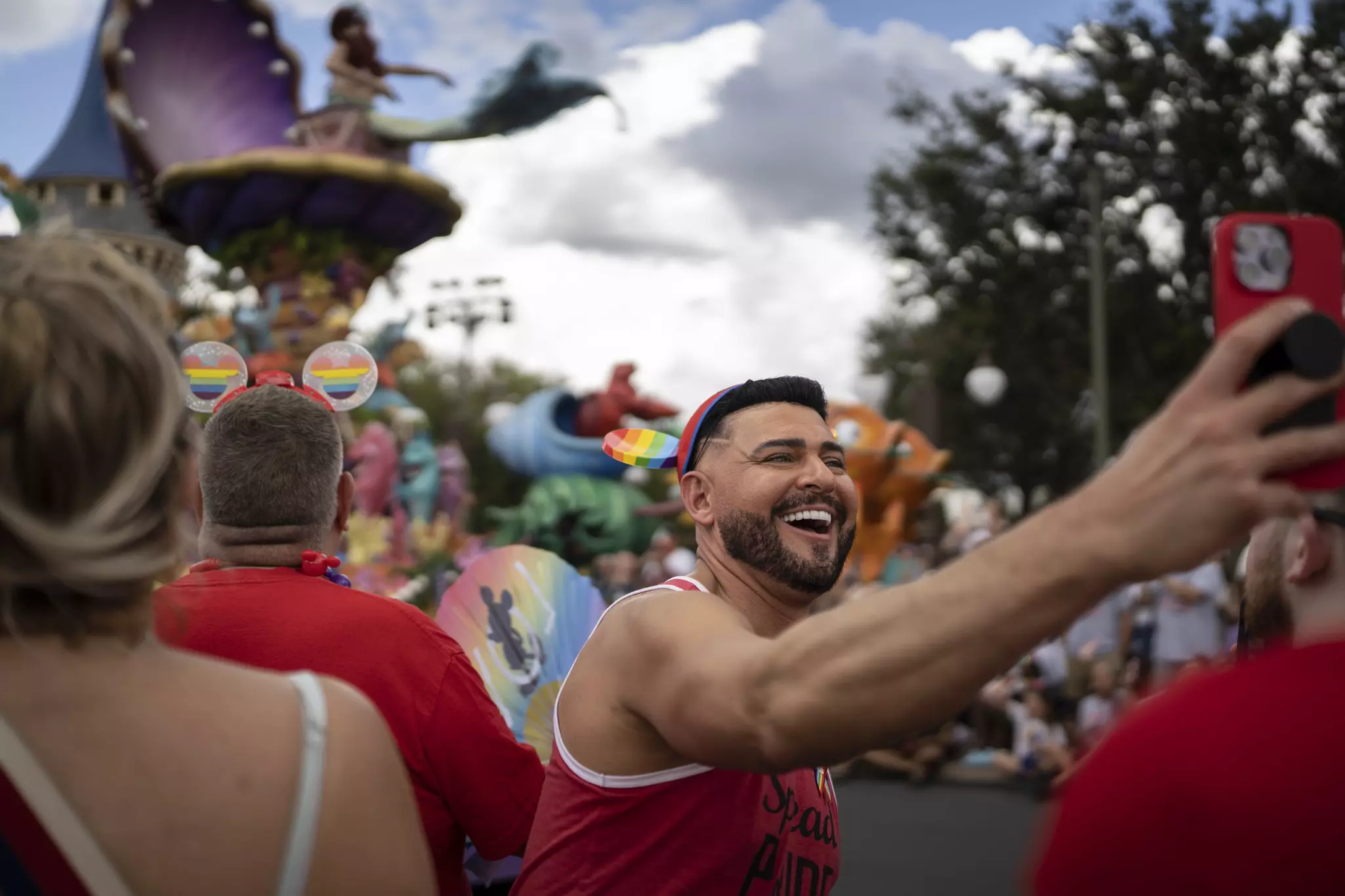 A smiling man taking a selfie in a theme park while wearing a red sleeveless shirt.
