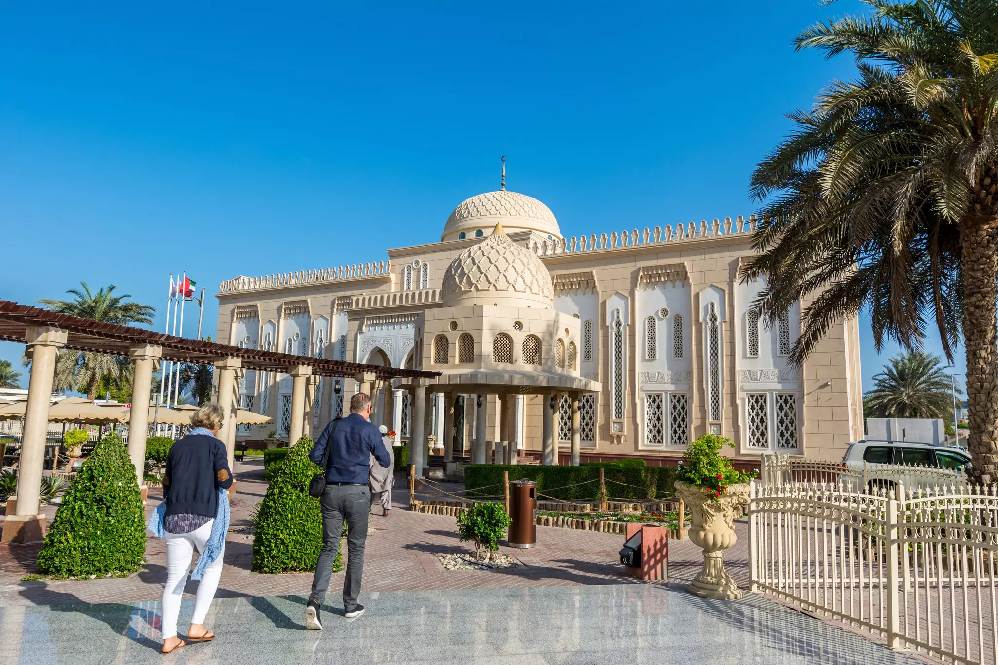 Two people walk toward a mosque in Dubai, United Arab Emirates, under clear blue sky.