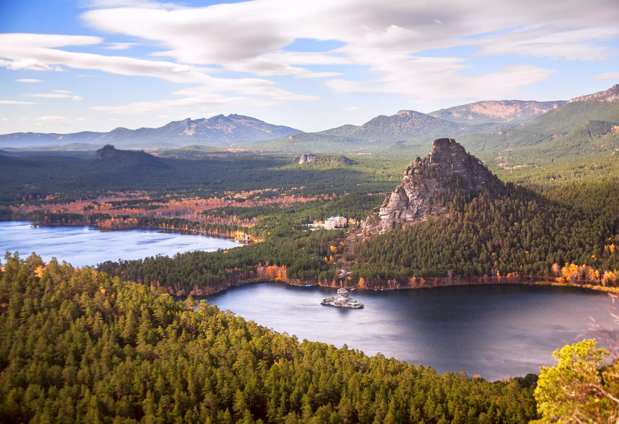 A wide view looking down at a lake surrounded by pine trees, a large rocky peak and other trees showing autumn foliage.