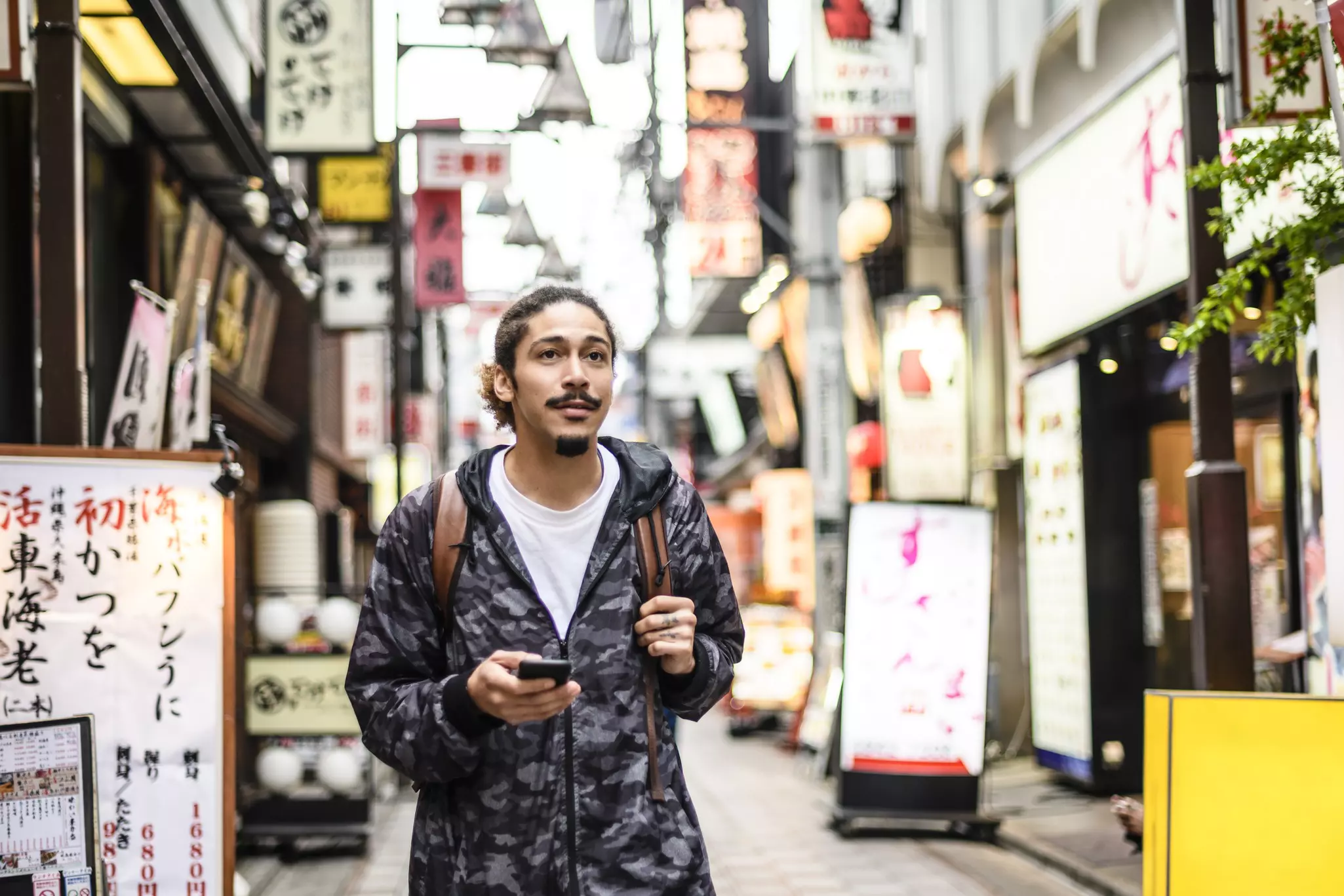A man holding a cell phone and carrying a backpack explores a street lined with signs in Japanese