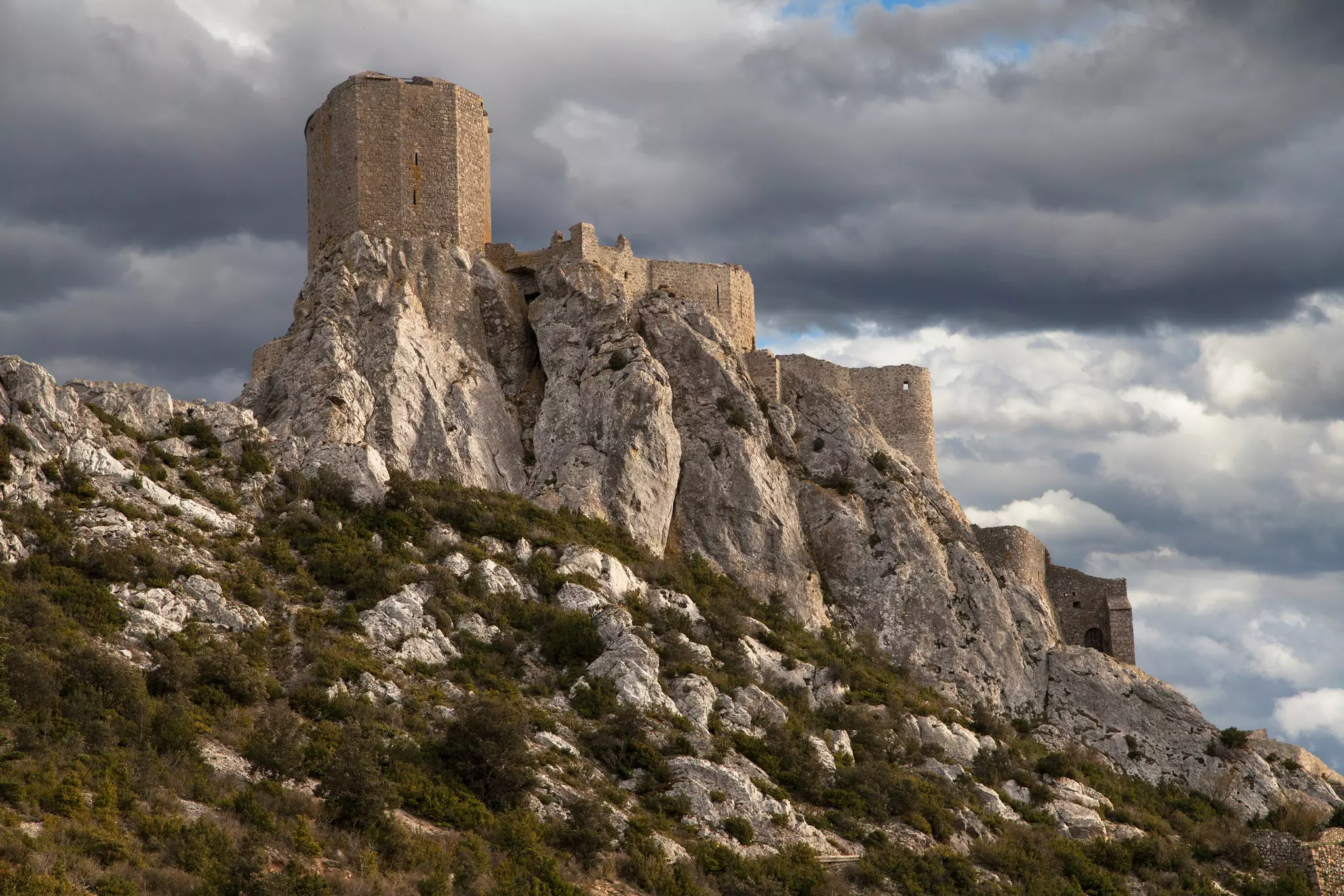 The Château de Quéribus rises over a rocky ridge against a dark sky in Languedoc, France. 