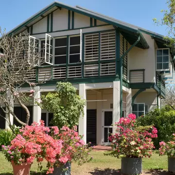 A two-storey house raised on piers with louvred verandah screens and green trim, bougainvillea in pots on the lawn in front.