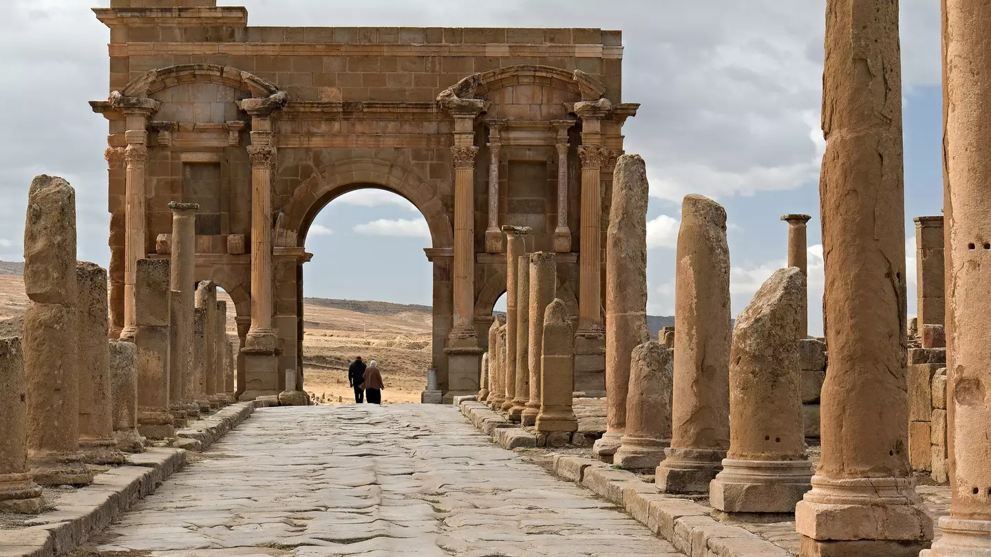 People walk under a large Roman archway in an archaeological site.