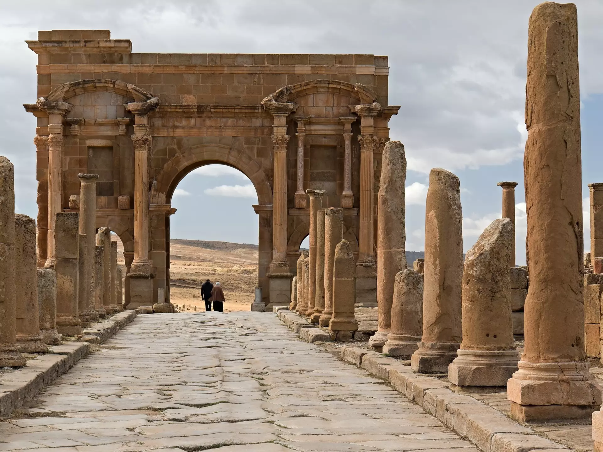 People walk under a large Roman archway in an archaeological site.