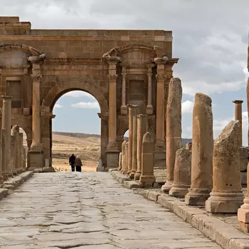 People walk under a large Roman archway in an archaeological site.