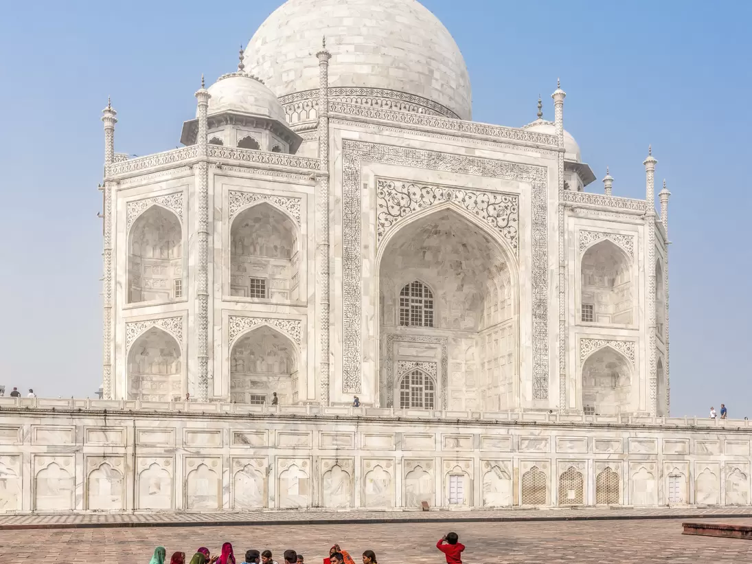 people relax around the walls of the Taj Mahal, a white building with domed roof and multiple spires. 