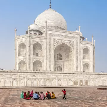 people relax around the walls of the Taj Mahal, a white building with domed roof and multiple spires. 