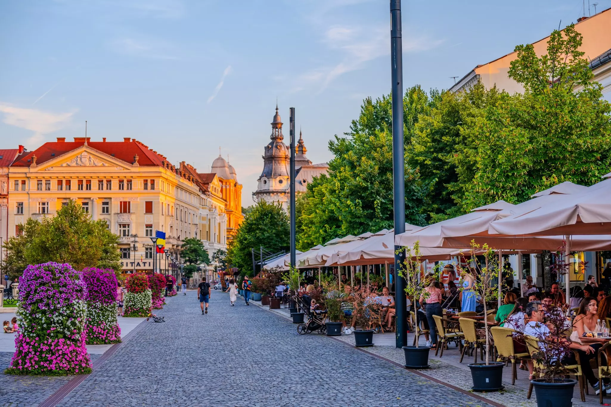 Dusk on a pedestrianized street lined with outdoor terraces where people are dining. The other side of the street is decorated with flowers, and children are playing.